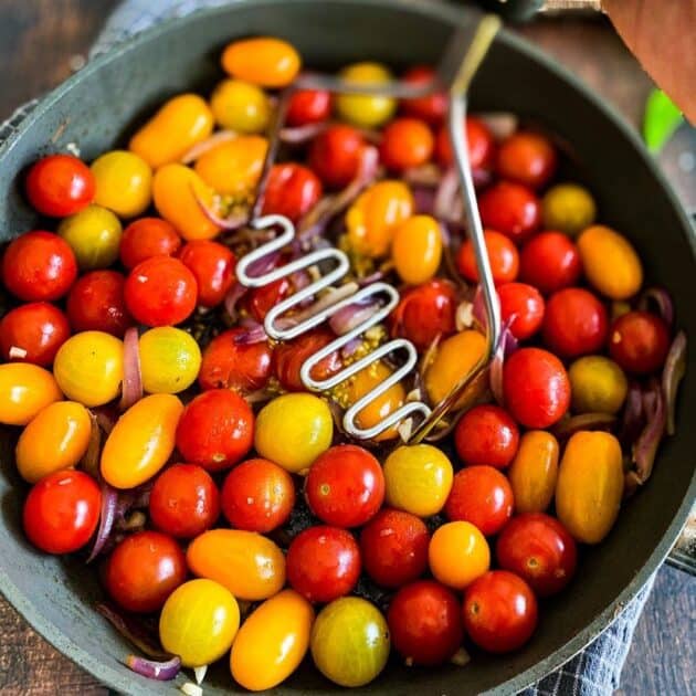 Potato masher crushing cherry tomatoes in a skillet.