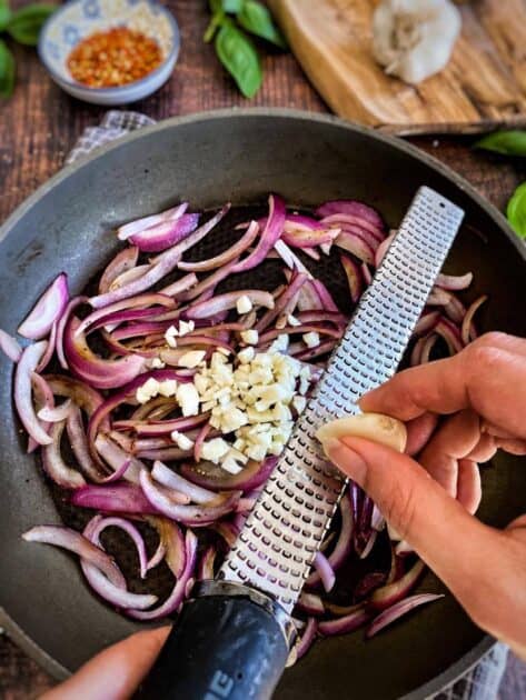 Hand grating garlic into a skillet with red onion.