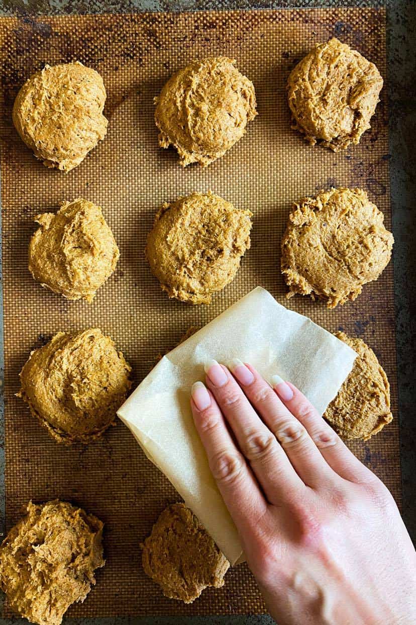 Vegan cookies on a baking sheet with a hand pressing them flat.