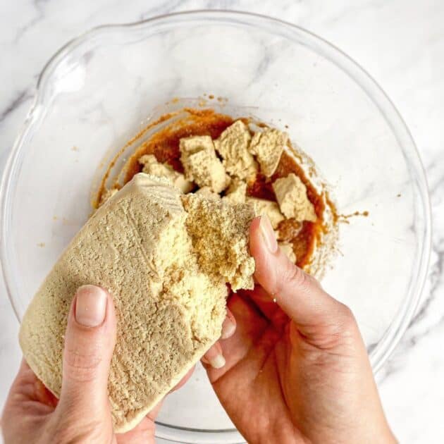 Hand breaking tofu nuggets into chunks over a bowl.