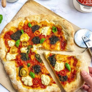 Vegan pesto pizza on a cutting board with a hand removing a slice.