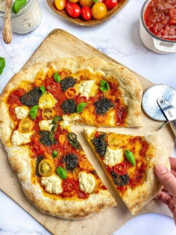 Vegan pesto pizza on a cutting board with a hand removing a slice.