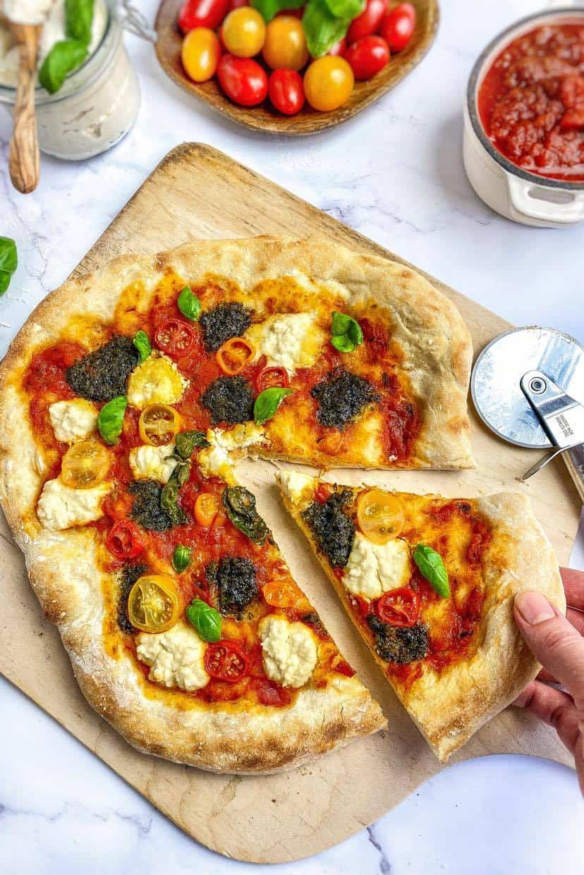 Vegan pesto pizza on a cutting board with a hand removing a slice.