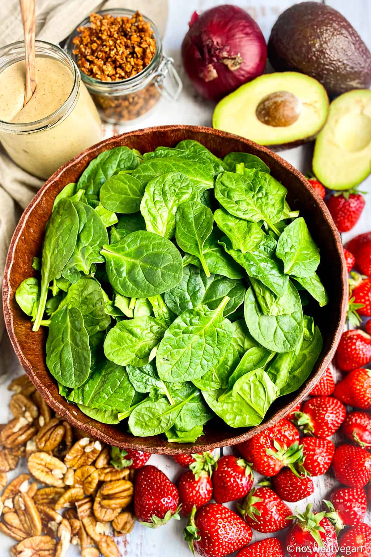 Ingredients for spinach salad with strawberries, avocado, red onion, pecans, vegan bacon bits, and creamy balsamic dressing.