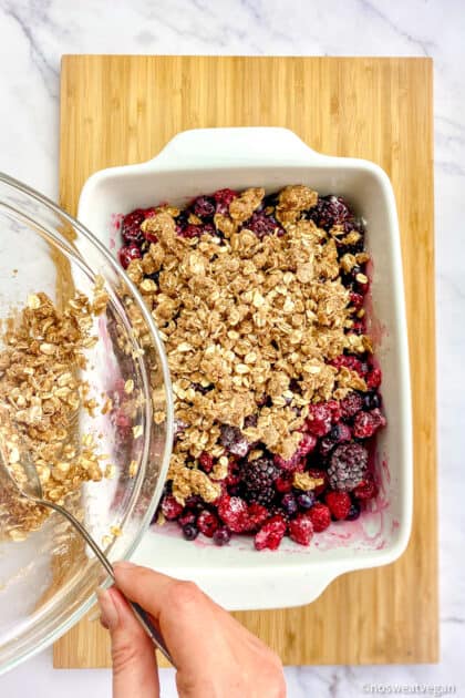 Hand spooning oat crumble topping over the frozen fruit mixture.
