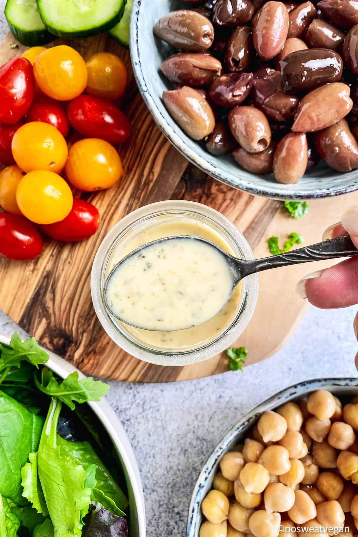 Greek buddha bowl dressing in center surrounded by tomatoes, olives, chickpeas, and mixed greens.