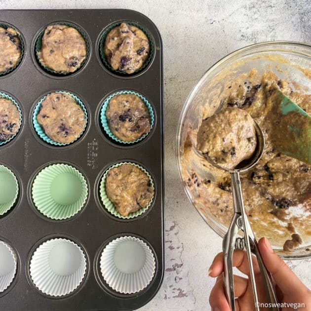 Hand using ice-cream scoop to transfer healthy muffin batter to cups.