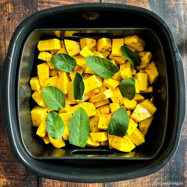 Uncooked butternut squash in air fryer basket with sage leaves.