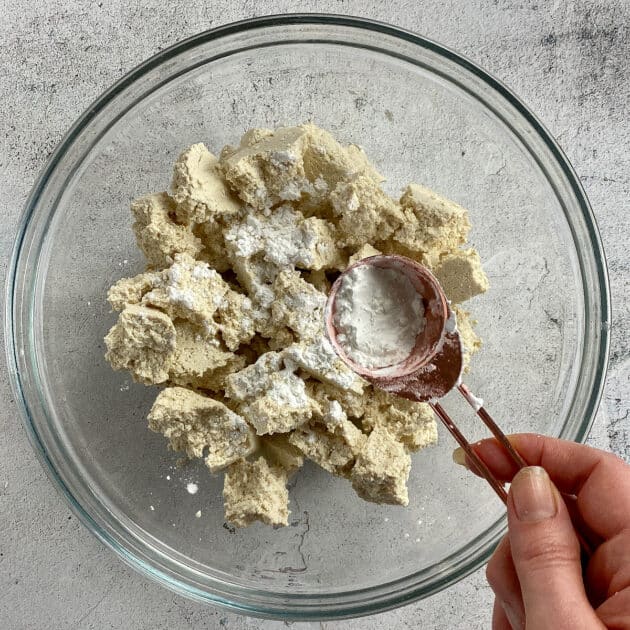 Hand adding cornstarch to bowl with tofu chunks.