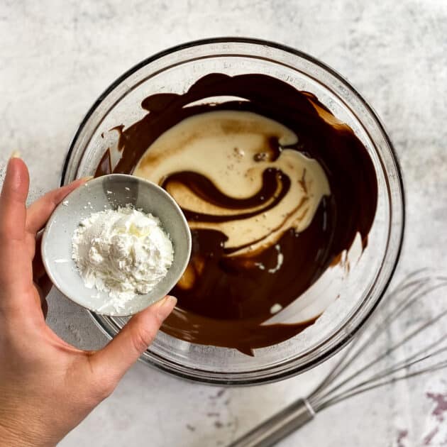 Hand adding cornstarch to bowl with melted chocolate and oat milk.