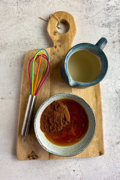 Cocoa powder and maple syrup in a bowl next to a whisk and small pitcher of water.