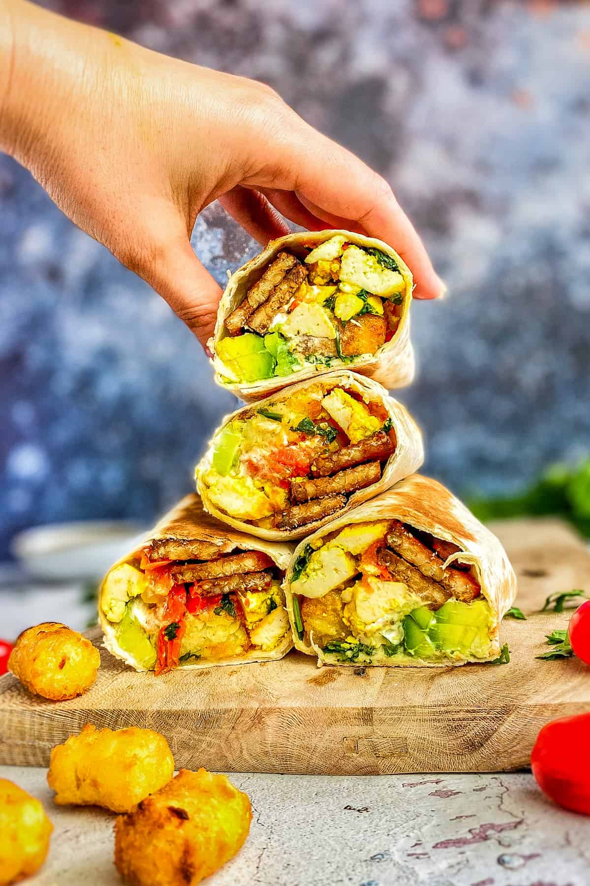 Vegan breakfast burritos stacked on a cutting board with hand taking one from the top.