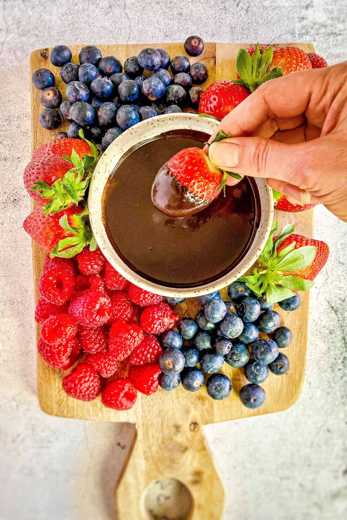 Hand dipping strawberry into a bowl of vegan chocolate fudge sauce surrounded by berries.