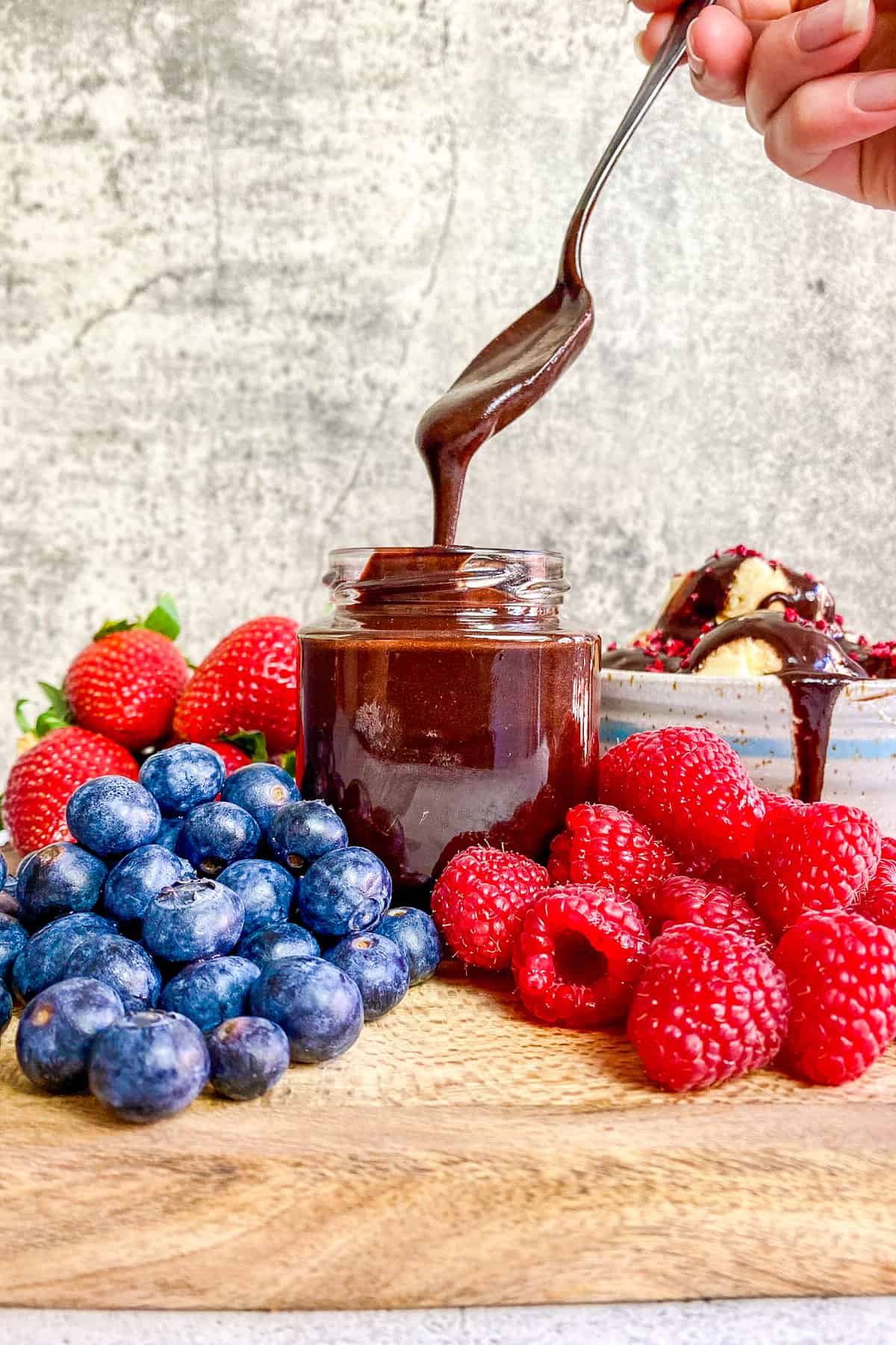 Hand lifting spoon of vegan chocolate sauce out of a jar surrounded by berries.