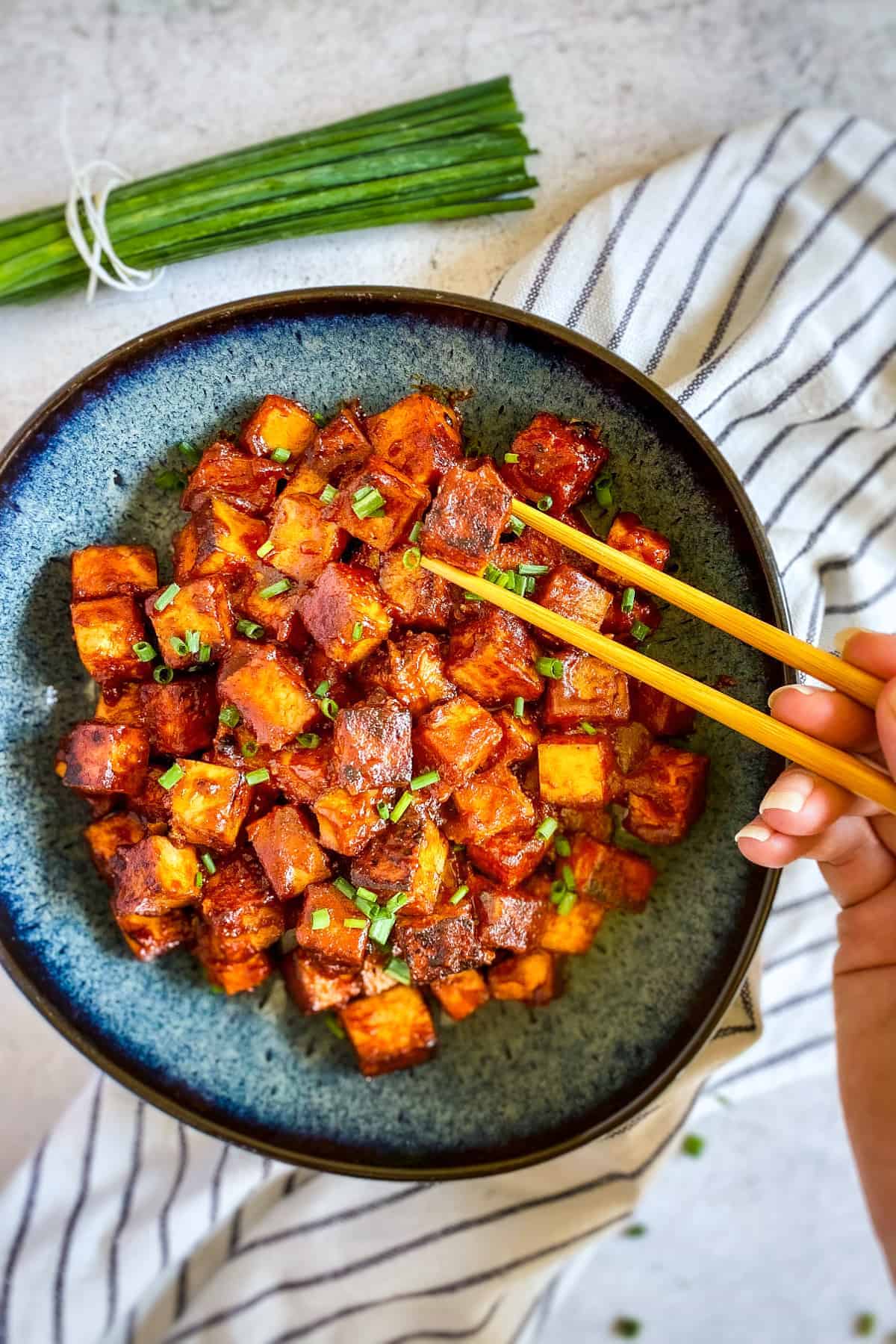Hand picking up a cube of marinated tofu with chopsticks.