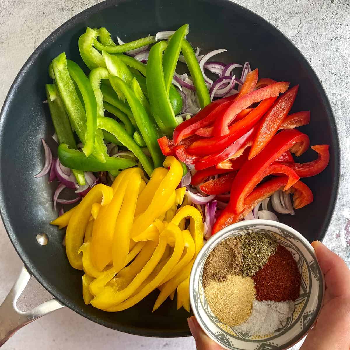 Peppers and onions in skillet with hand hold bowl of fajita veggie seasoning.