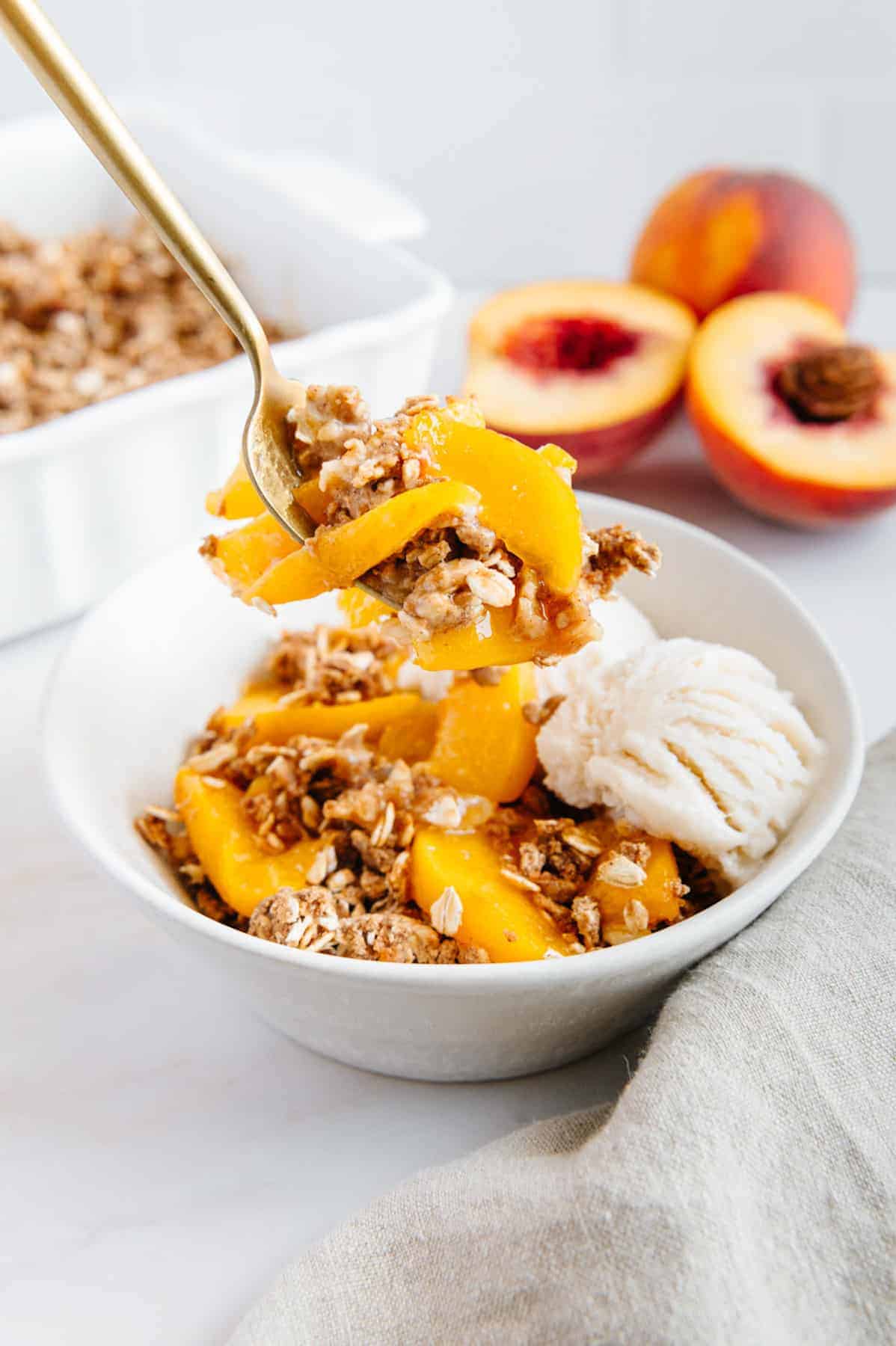Vegan peach crisp in a bowl with ice cream and a spoon lifting a bite out.