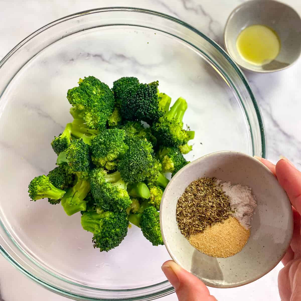 Broccoli in bowl with hand holding seasonings.