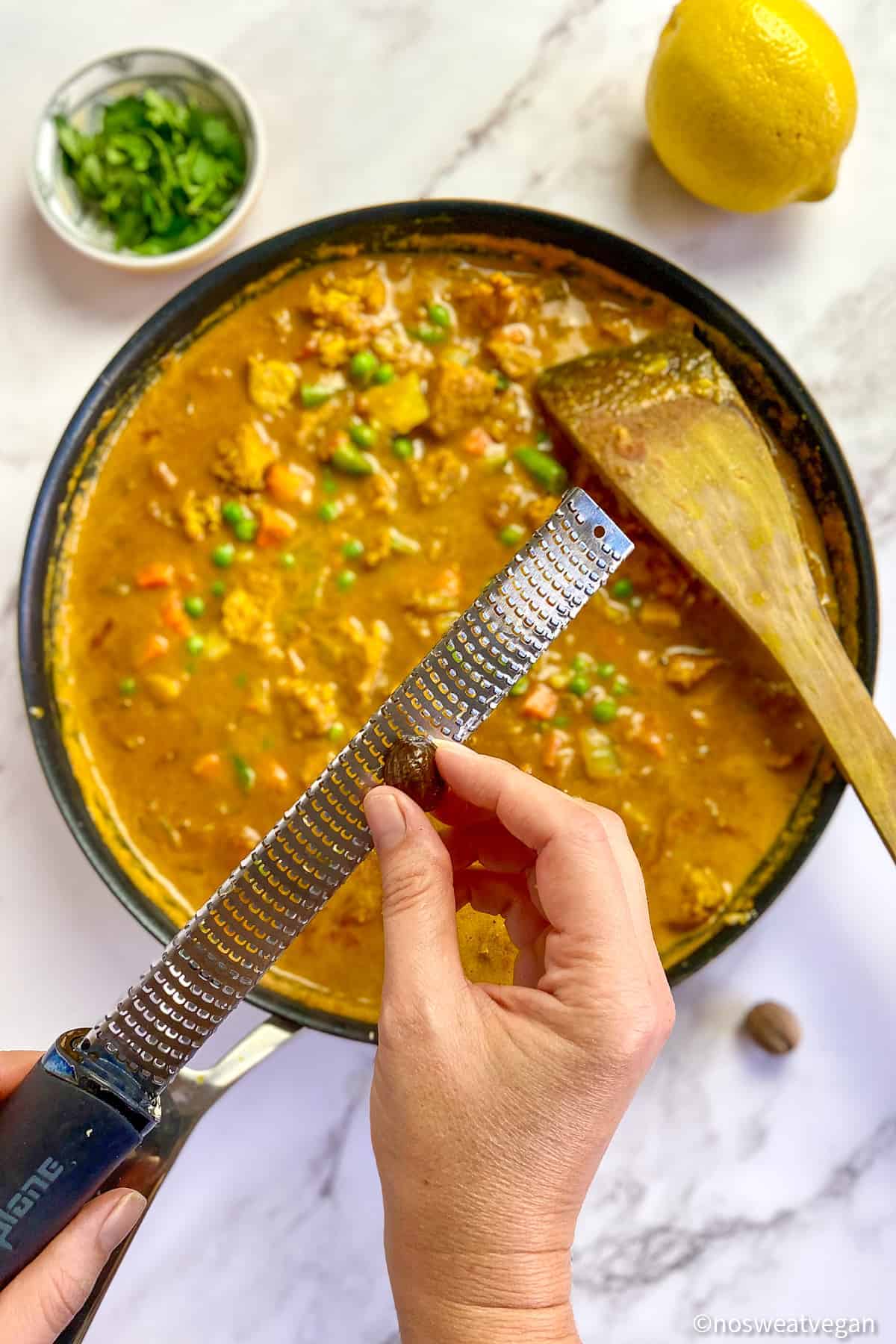 Hands grating nutmeg over a skillet with vegan curry.