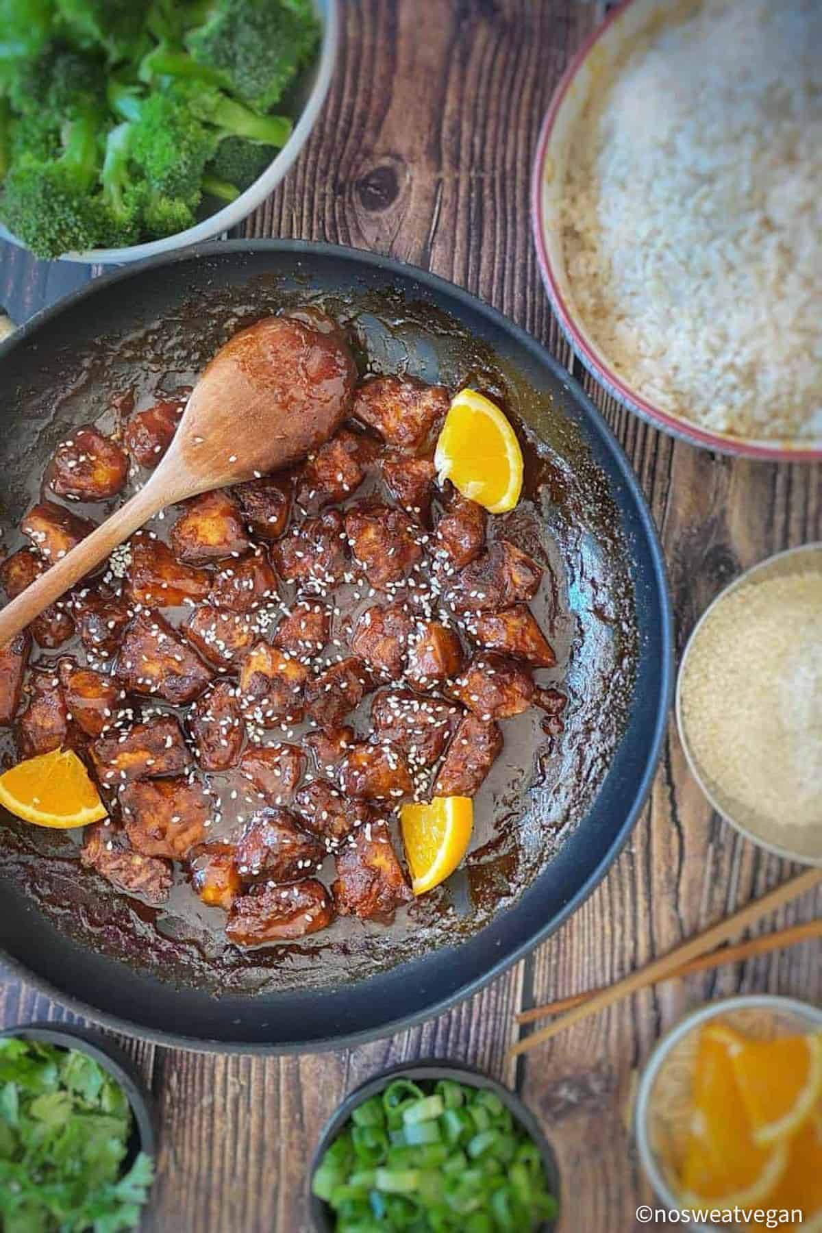Vegan orange chicken in skillet surrounded by broccoli, rice, and garnishes.