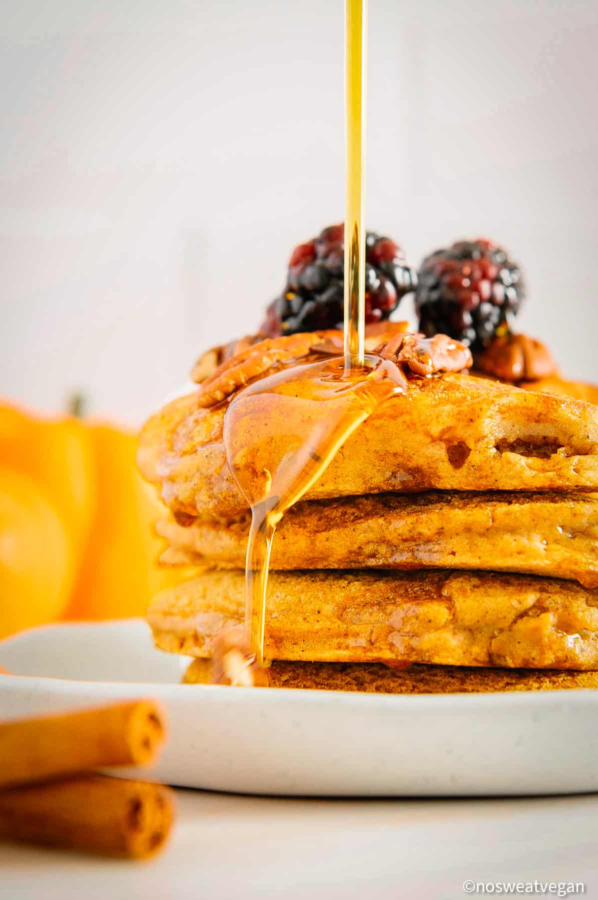Pumpkin pancakes stacked on a plate with maple syrup pouring over the top.