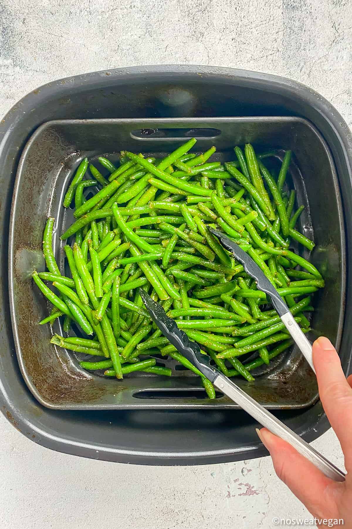 Hand using tongs to transfer frozen green beans into air fryer.