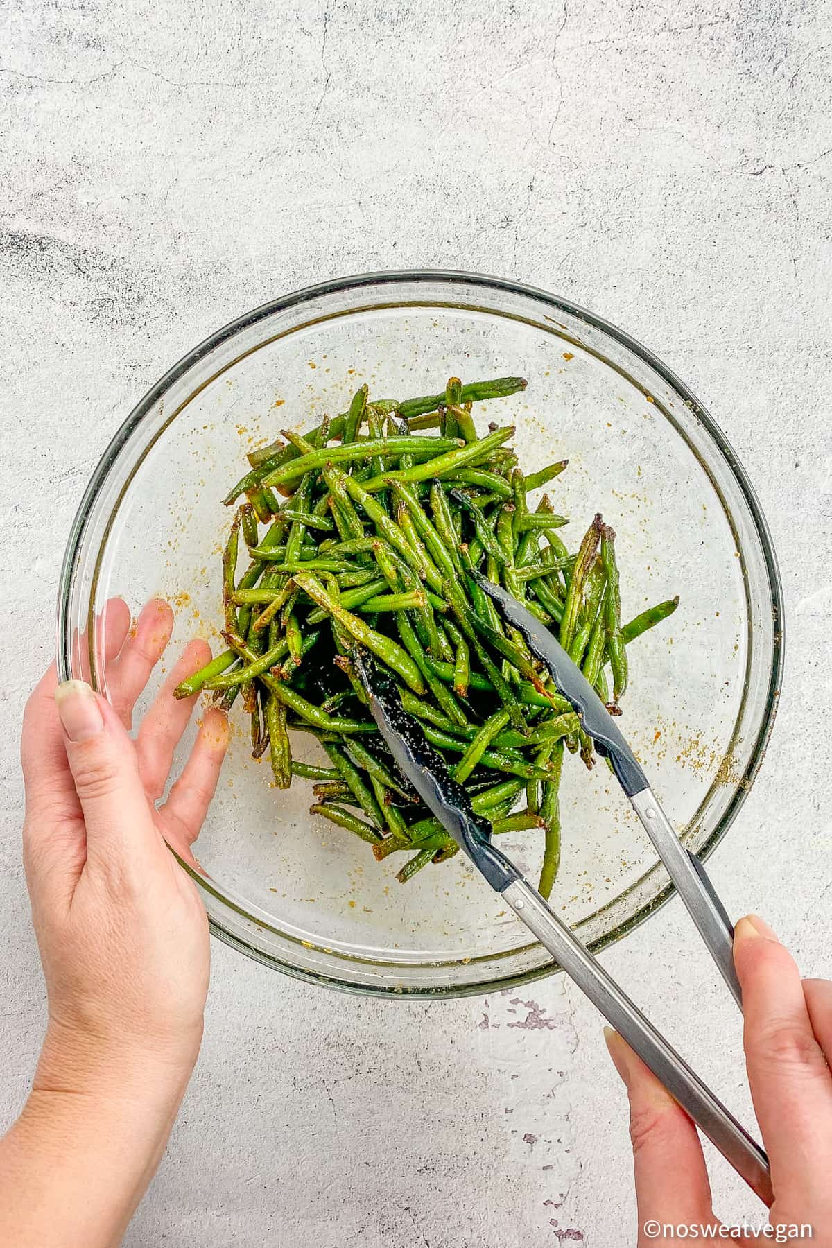 Air fried frozen green beans in a bowl with two hands mixing the green beans with tongs.