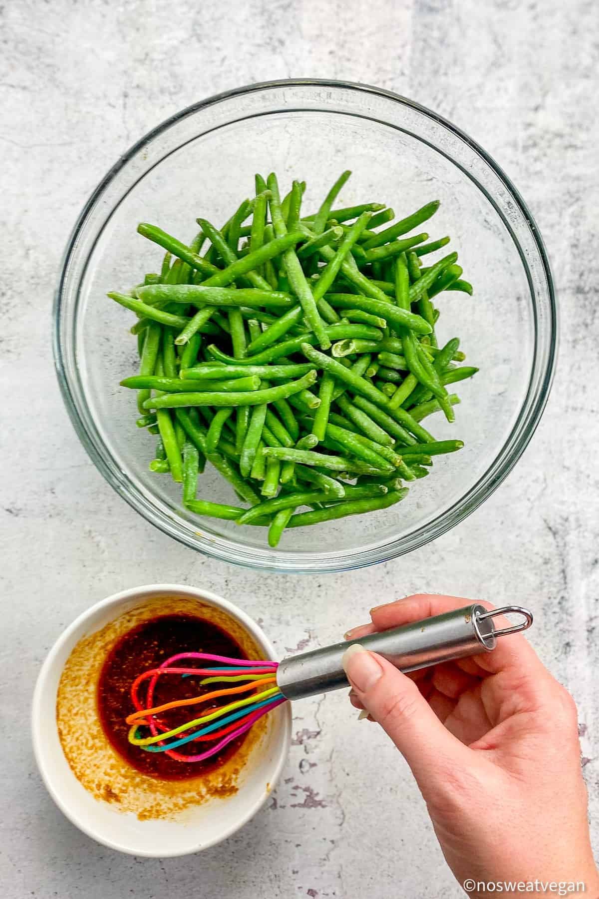Frozen green beans in a large bowl next to a small bowl with a hand whisking seasoning.