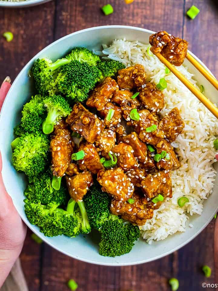 Bowl with teriyaki tofu, rice, and broccoli. Hand using chopsticks to pick up a chunk of tofu.