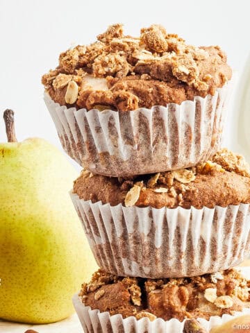 Three vegan pear muffins stacked in front of a pear.