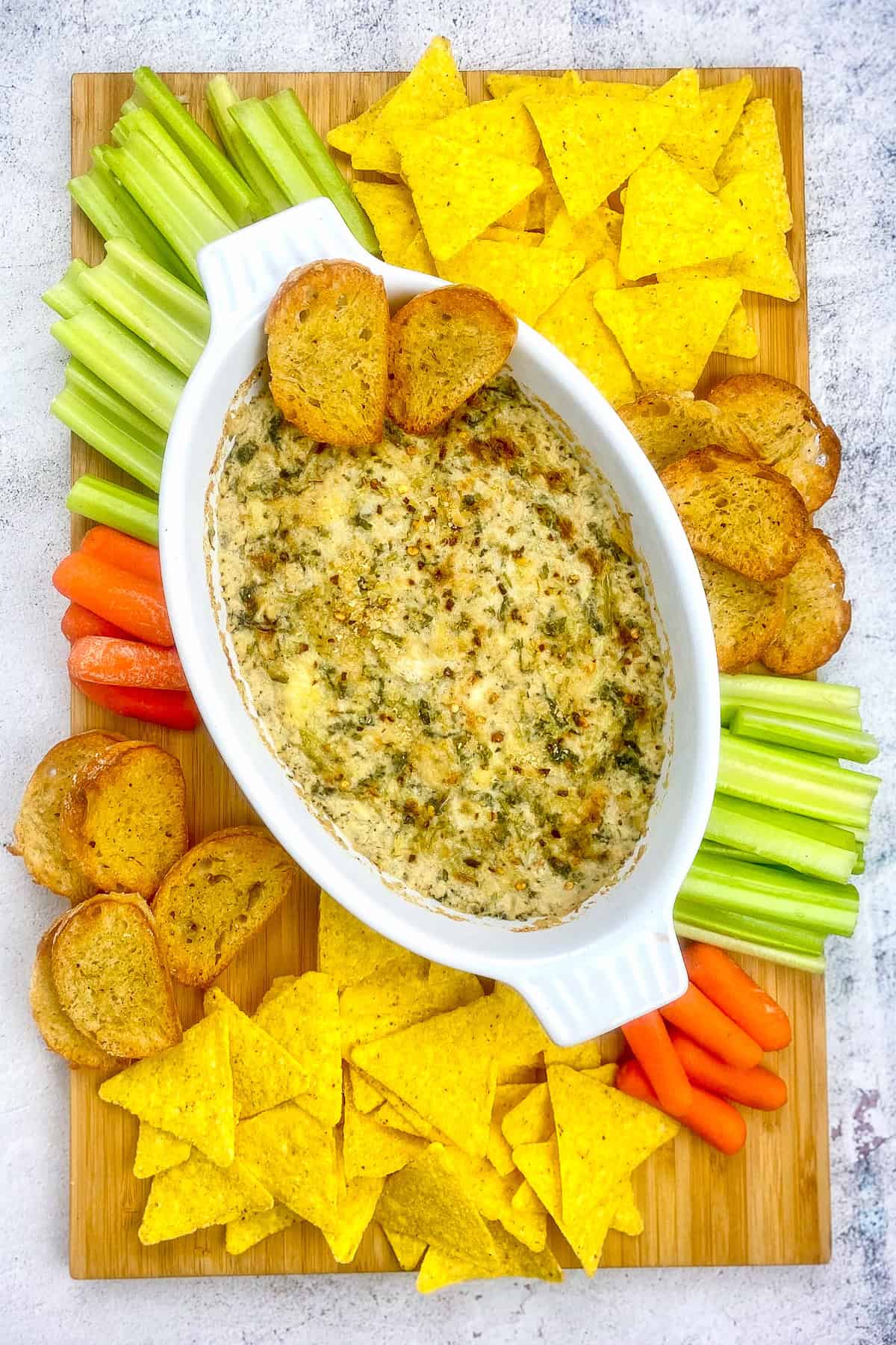 Vegan spinach artichoke dip on a cutting board with chips, toast, celery, and carrots.